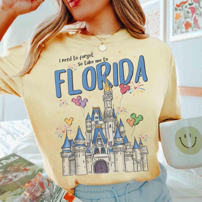 May include: Pale yellow t-shirt with the words "I need to forget so take me to FLORIDA" in blue. The shirt features a castle graphic with Mickey Mouse balloons and fireworks. A white mug with a smiley face is held in the hand.
