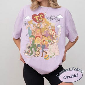 May include: A light purple t-shirt featuring a large graphic print of various animated characters in a collage style. The design includes a red heart with the letters TS, musical notes, clouds, and a colorful ball. The shirt has a crew neckline.