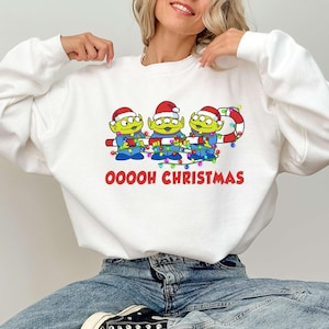 Könnte beinhalten: Weißes Sweatshirt mit einer Grafik von drei grünen Aliens mit Weihnachtsmützen, die Lichterketten halten. Der rote Text "Ooooh Christmas" ist darunter gedruckt. Ein festliches Weihnachtsdesign.
