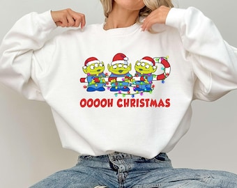 May include: White sweatshirt featuring a graphic of three green aliens wearing Santa hats and holding Christmas lights. The text "Ooooh Christmas" is printed in red below the aliens. The sweatshirt is a festive holiday design.