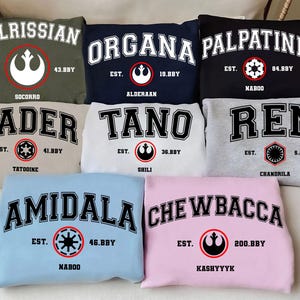 Op de afbeelding: Een verzameling sweatshirts in verschillende kleuren, elk met een naam in vette, gebogen letters. De namen zijn "Calrissian", "Organa", "Palpatine", "Vader", "Tano", "Ren", "Amidala" en "Chewbacca". Elk sweatshirt toont ook een symbool en de tekst "EST." met een nummer en een locatie.