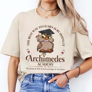 May include: A beige t-shirt with a brown graphic of an owl wearing a graduation cap. The graphic is surrounded by a circle with the text "The best way to learn is by doing it". Below the graphic is the text "Archimedes Academy of Wisdom & Wit & Knowledge & Sarcasm".