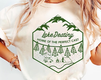 May include: Cream-colored sweatshirt with a green graphic. The graphic features the text "Lake Destiny" above a camping scene with trees, mountains, a car, a tent, and a campfire. The phrase "HOME OF THE PERFECT CAST" is also included.