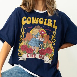 K&ouml;nnte beinhalten: Ein marineblaues T-Shirt mit einem Grafikdruck und dem Text COWGIRL LIKE ME. Das Design zeigt zwei Figuren in Westernkleidung in einer W&uuml;stenlandschaft mit Kakteen, roten Rosen und einem Sonnenuntergang, umrahmt von gelben dekorativen Schn&ouml;rkeln.