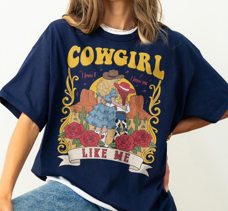 K&ouml;nnte beinhalten: Ein marineblaues T-Shirt mit einem Grafikdruck und dem Text COWGIRL LIKE ME. Das Design zeigt zwei Figuren in Westernkleidung in einer W&uuml;stenlandschaft mit Kakteen, roten Rosen und einem Sonnenuntergang, umrahmt von gelben dekorativen Schn&ouml;rkeln.