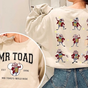 Könnte beinhalten: Ein beiger Pullover mit einem grafischen Aufdruck von Mr. Toad aus Disneys The Wind in the Willows. Der Druck enthält den Text "Mr. Toad", "Est. 1996" und "Mr. Toad's Wild Ride".