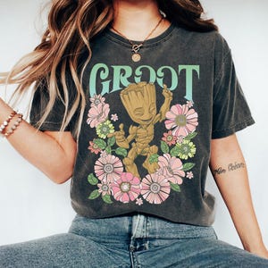 May include: Dark gray t-shirt featuring the word "GROOT" in a retro font, with a cartoon Groot character surrounded by pink and yellow flowers. The shirt has a vintage, faded look.