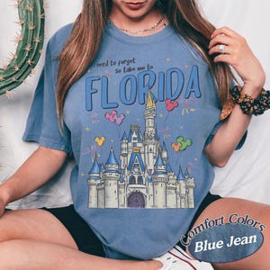 May include: Blue jean coloured t-shirt featuring a castle graphic, balloons, and fireworks. The text reads "I need to forget so take me to FLORIDA". The shirt is a Comfort Colors brand.