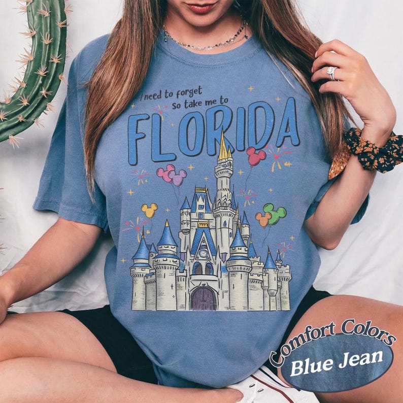 May include: Blue jean coloured t-shirt featuring a castle graphic, balloons, and fireworks. The text reads "I need to forget so take me to FLORIDA". The shirt is a Comfort Colors brand.