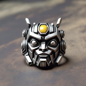 Könnte beinhalten: Ein silberfarbener Ring in Form eines Roboterkopfes. Der Ring hat detaillierte Merkmale, darunter Augen, einen Mund und einen gelben Edelstein oben. Der Ring besteht aus Metall und hat ein detailliertes, mechanisches Design.