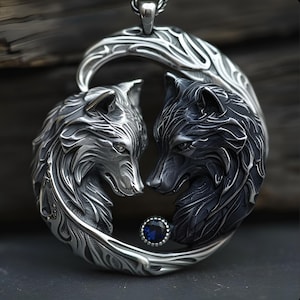 May include: A circular silver pendant featuring two sculpted wolf heads facing each other in a yin-yang formation. One wolf is polished silver, the other is oxidized dark grey. A small, round, faceted blue gemstone is set between the two wolf heads.