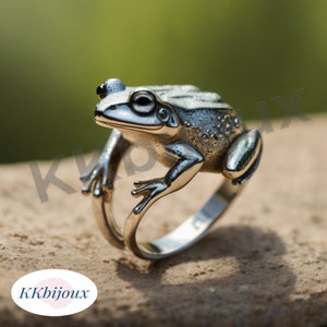 Könnte beinhalten: Ein silberfarbener Metallring mit einer detaillierten, dreidimensionalen Froschfigur auf der Oberseite. Der Frosch hat eine texturierte Haut, hervorstehende Augen und ausgestreckte Beine, die den Ring bilden. Der Text KKbijoux ist zu sehen.