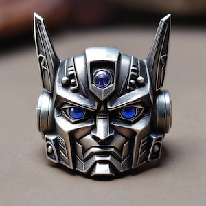 Op de afbeelding: Een zilverkleurige metalen ring in de vorm van een robotkop met puntige oren. Het ontwerp heeft een donkere, geborstelde afwerking met ingewikkelde geometrische details en drie ingebedde ronde blauwe edelstenen op het voorhoofd en als ogen.