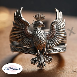 May include: A silver-toned ring featuring a detailed phoenix with outstretched wings and a crest on its head. The bird is centered on a band with an intricate braided pattern. Small brown stones are embedded along the wings. Text reads KKbijoux.