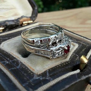 Op de afbeelding: Een zilveren ringenset met een vierkante robijn en meerdere kleine diamanten, rustend in een vintage sieradendoosje. De ring heeft een gedetailleerd ontwerp met ronde en baguette-geslepen diamanten. De doos is donker met een fluwelen binnenkant.