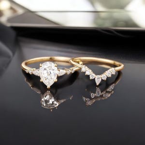 May include: A pear-shaped diamond engagement ring with a delicate band and a matching pear-shaped diamond wedding band. Both rings are set in gold and feature small diamonds.
