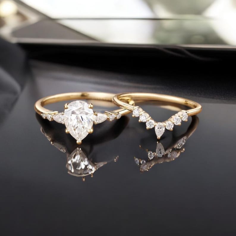 May include: A pear-shaped diamond engagement ring with a delicate band and a matching pear-shaped diamond wedding band. Both rings are set in gold and feature small diamonds.