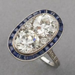 May include: A silver ring with two oval-shaped diamonds set in a bezel setting. The diamonds are surrounded by a halo of blue sapphires.