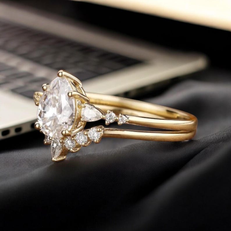 May include: A gold engagement ring with a pear-shaped centre stone and smaller stones set around it. The ring is on a grey fabric background.
