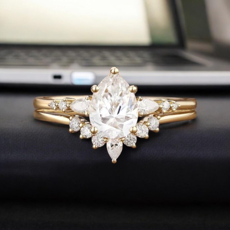 May include: A pear-shaped diamond engagement ring set in yellow gold with a halo of smaller diamonds. The ring is on a black surface.