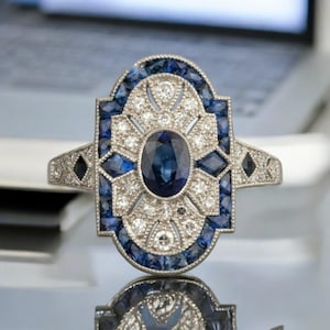 Peut inclure: Bague de style Art Déco en argent, ornée d'un saphir ovale central, entouré de petits saphirs bleus et de diamants blancs étincelants. Le design géométrique de la bague reflète la lumière.