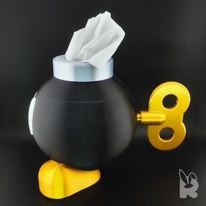 Side view of Super Mario Bob-omb tissue box.  Gold feet, black body, white eyes, and silver cap with tissue protruding from the top to form the wick