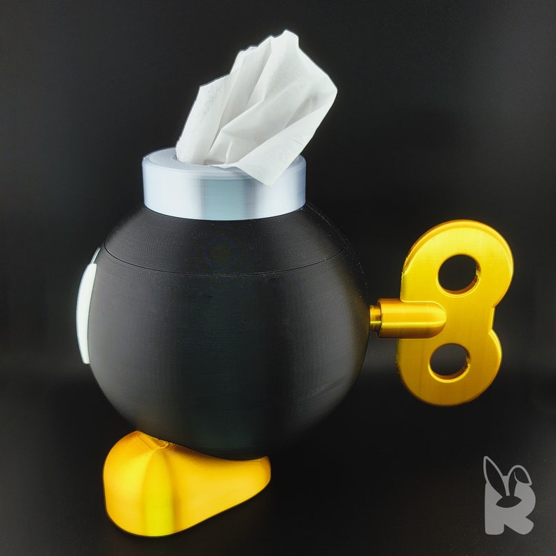 Side view of Super Mario Bob-omb tissue box.  Gold feet, black body, white eyes, and silver cap with tissue protruding from the top to form the wick