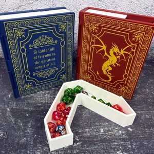 May include: Two book-shaped dice boxes, one blue with gold accents and text, the other red with a gold dragon design. A white dice tray holds various colored dice. The blue box reads: "A table full of friends is the greatest magic of all."