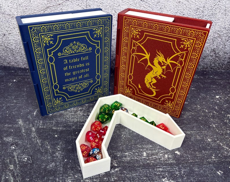 May include: Two book-shaped dice boxes, one blue with gold accents and text, the other red with a gold dragon design. A white dice tray holds various colored dice. The blue box reads: "A table full of friends is the greatest magic of all."