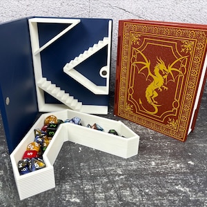 May include: A navy blue dice tower with white stairs and a white dice tray filled with colorful dice. A red book with a gold dragon design is next to the tower. The items are on a gray surface.
