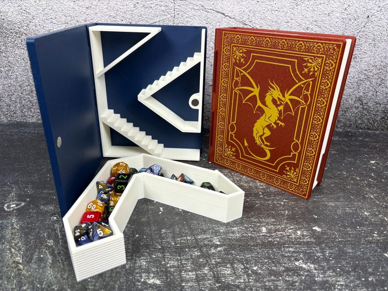 May include: A navy blue dice tower with white stairs and a white dice tray filled with colorful dice. A red book with a gold dragon design is next to the tower. The items are on a gray surface.
