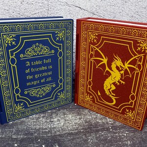 May include: Two decorative books with ornate covers. One is navy blue with gold accents and the quote, "A table full of friends is the greatest magic of all." The other is burgundy with a gold dragon design.