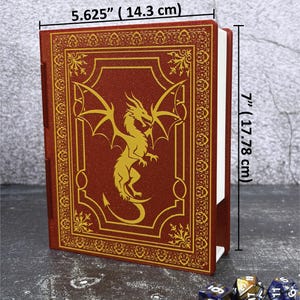 May include: A red, faux-leather book with a gold dragon design and decorative border. The book measures 5.625 inches (14.3 cm) wide and 7 inches (17.78 cm) tall. The book is likely for role-playing games.