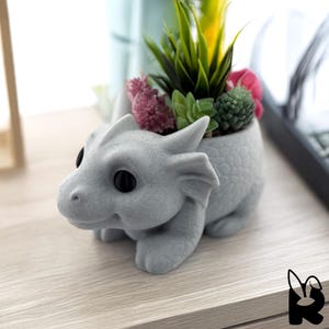 Op de afbeelding: Een grijze draakvormige plantenbak met zwarte ogen, gevuld met diverse kunstmatige vetplanten en bloemen. De plantenbak is gemaakt van een textuurrijk materiaal en staat op een lichtgekleurd houten oppervlak.