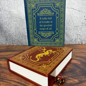 May include: Two decorative book-shaped boxes. One is blue with gold filigree and the text "A table full of friends is the greatest magic of all." The other is maroon with a gold dragon design. A red and black dice is next to the maroon box.