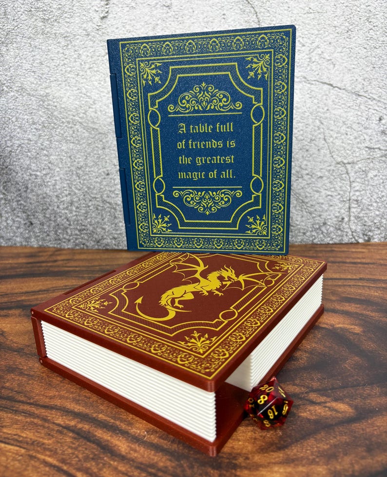 May include: Two decorative book-shaped boxes. One is blue with gold filigree and the text "A table full of friends is the greatest magic of all." The other is maroon with a gold dragon design. A red and black dice is next to the maroon box.
