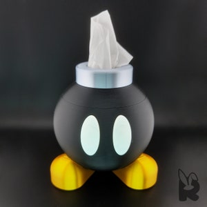 Front view of Super Mario Bob-omb tissue box. Gold feet, black body, white eyes, and silver cap with tissue protruding from the top to form the wick.
