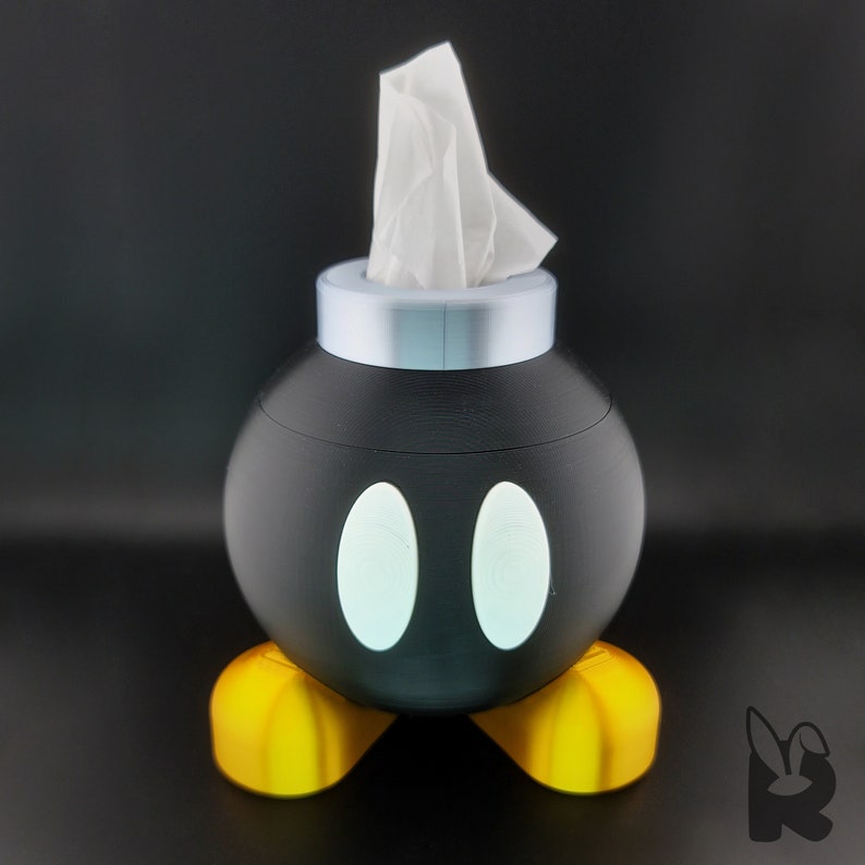 Front view of Super Mario Bob-omb tissue box. Gold feet, black body, white eyes, and silver cap with tissue protruding from the top to form the wick.