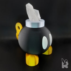 Isometric view Super Mario Bob-omb tissue box.  Gold feet, black body, white eyes, and silver cap with tissue protruding from the top to form the wick