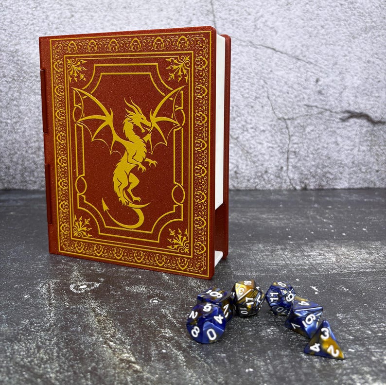 May include: A red, faux-leather book-shaped dice box with a gold dragon design on the front. The box is open, revealing white pages. Several multi-sided dice in blue and gold are scattered in front of the box.