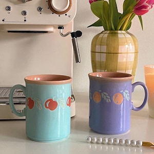 May include: Two ceramic mugs with fruit designs. The mug on the left is light blue with a red cherry design. The mug on the right is purple with an orange design. Both mugs have a pink rim.