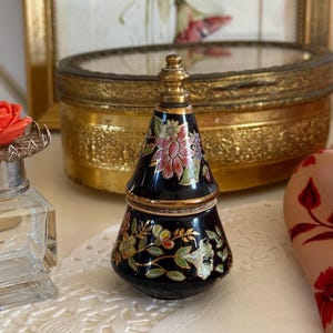 Può includere: Un piccolo vasetto conico in ceramica nera con un coperchio metallico dorato. L'esterno presenta un motivo floreale dipinto a mano nei toni del rosa, verde e bianco con contorni dorati. Il vasetto ha una base larga che si restringe verso l'alto.