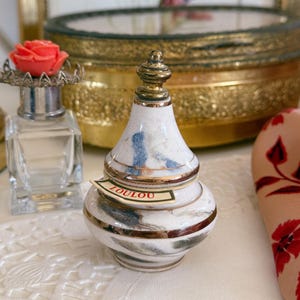 Può includere: Un piccolo flacone di profumo in ceramica a forma di pera con base bianca, motivi marmorizzati blu e grigi e finiture in metallo dorato. Il flacone presenta un tappo in metallo color oro e una piccola etichetta di carta con la scritta LOULOU in rosso.