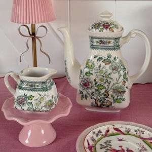 Puede incluir: Una pantalla de lámpara plisada rosa sobre una base de lámpara dorada, una tetera blanca con diseños florales, una pequeña crema en un soporte para pasteles rosa y un plato decorativo con ilustraciones de aves. Los artículos están dispuestos sobre un mantel rosa.