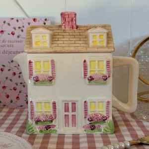 Peut inclure: Une théière en céramique blanche en forme de maison, avec un toit beige et une cheminée rose. La maison a des fenêtres jaunes avec des volets roses et des jardinières. Une nappe à carreaux roses et blancs est au premier plan.