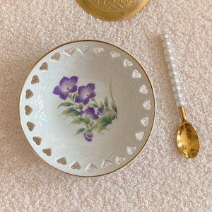 Könnte beinhalten: Eine weiße Porzellanschale mit goldfarbenem Rand und einem Blumendesign. Die Schale hat einen gewellten Rand mit herzförmigen Ausschnitten. Ein goldener Löffel mit einem perlmuttartigen Griff liegt neben der Schale.