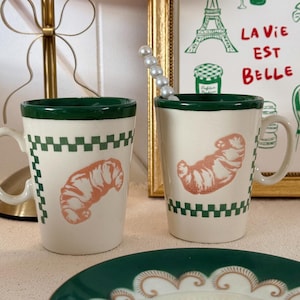 Op de afbeelding: Twee off-white keramische mokken met een donkergroene rand en een groen geblokt patroon. Elke mok is voorzien van een perzikkleurige croissantillustratie. Eén mok bevat een decoratieve roerstaaf met witte bolvormige kralen aan de bovenkant.