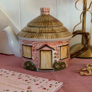 Può includere: Teiera in ceramica a forma di casetta, con tetto di paglia e pareti a mattoni rosa e marroni. Il beccuccio è bianco e il manico marrone. Dettagli floreali vicino alla porta e alle finestre.