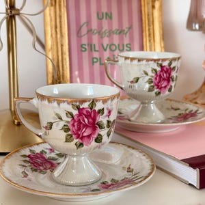 May include: Two white porcelain teacups and matching saucers featuring a pink rose floral pattern, iridescent finish, and gold-painted scalloped rims. Each cup is supported by a pedestal base, and the saucers display a coordinating rose design.
