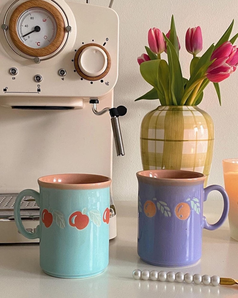 May include: Two ceramic mugs with fruit designs. The mug on the left is light blue with a red cherry design. The mug on the right is purple with an orange design. Both mugs have a pink rim.